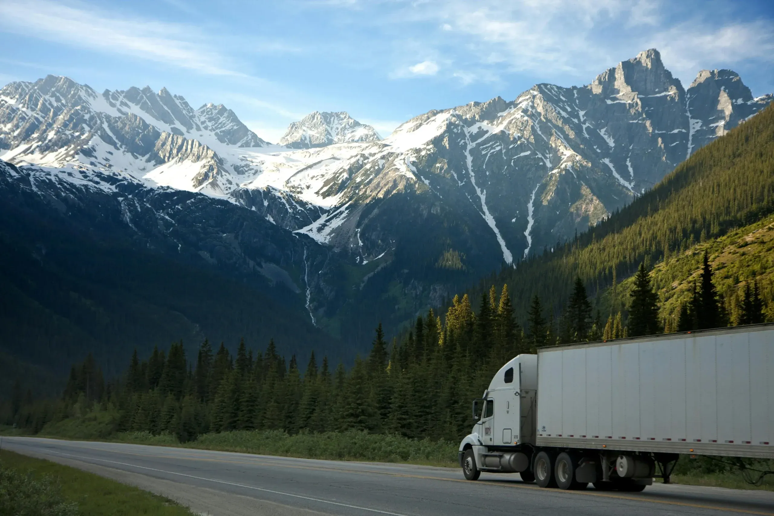 Ein weißer Lkw fährt auf einer Autobahn entlang, im Hintergrund schneebedeckte Berge und Kiefernwälder unter einem strahlend blauen Himmel.
