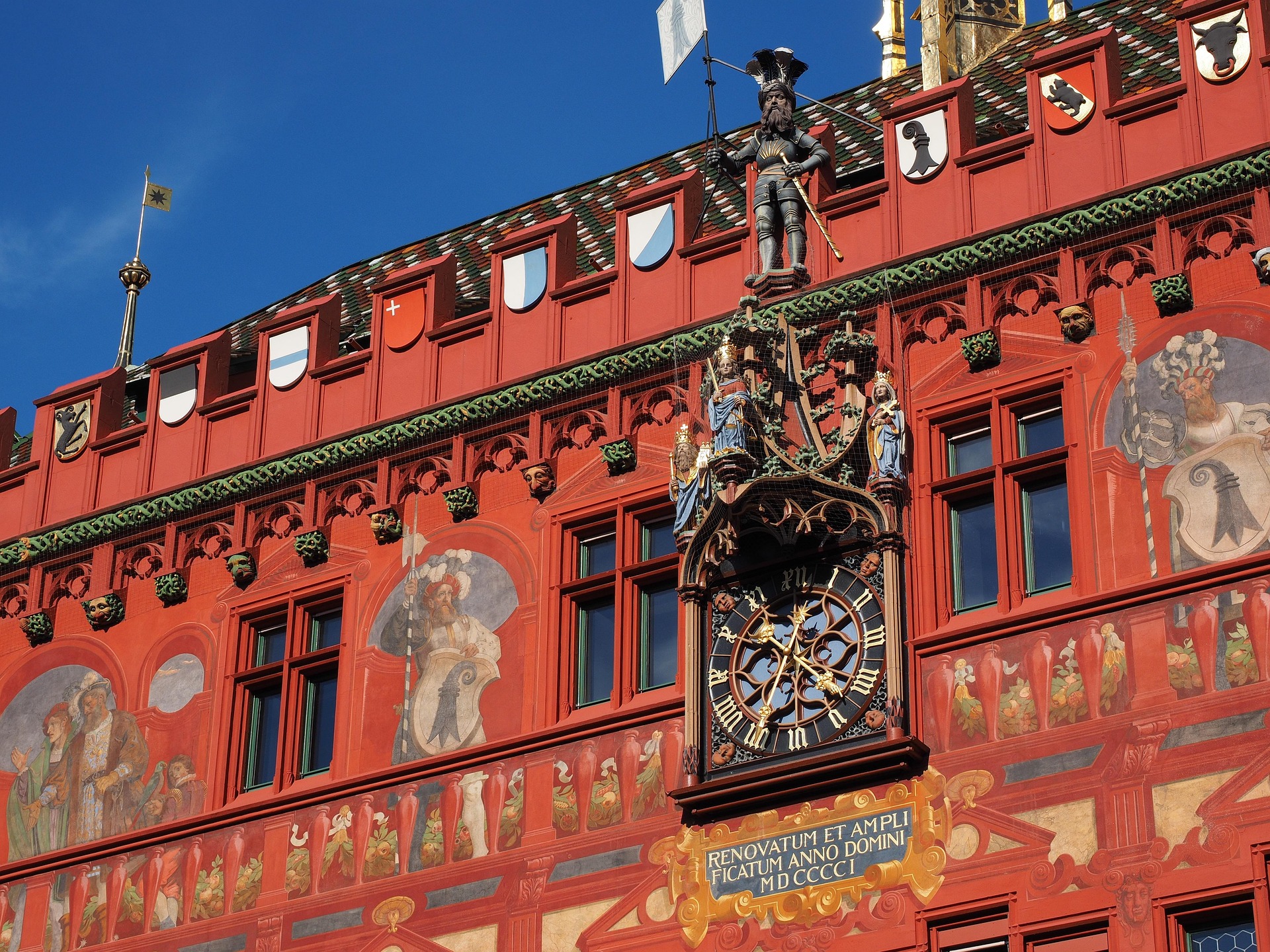 Detailansicht des roten Basler Rathauses mit kunstvollen Fassadenmalereien, Uhr und historischen Figuren unter blauem Himmel.