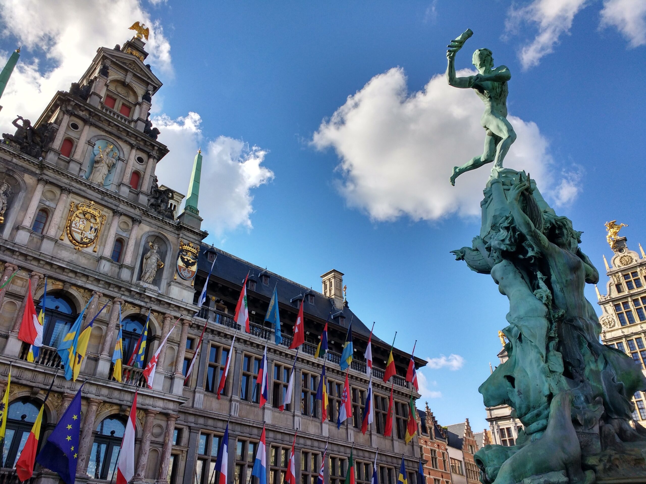 Historisches Rathaus und Brabobrunnen auf dem Grote Markt in Antwerpen, Belgien, unter blauem Himmel mit internationalen Flaggen und barocker Architektur.