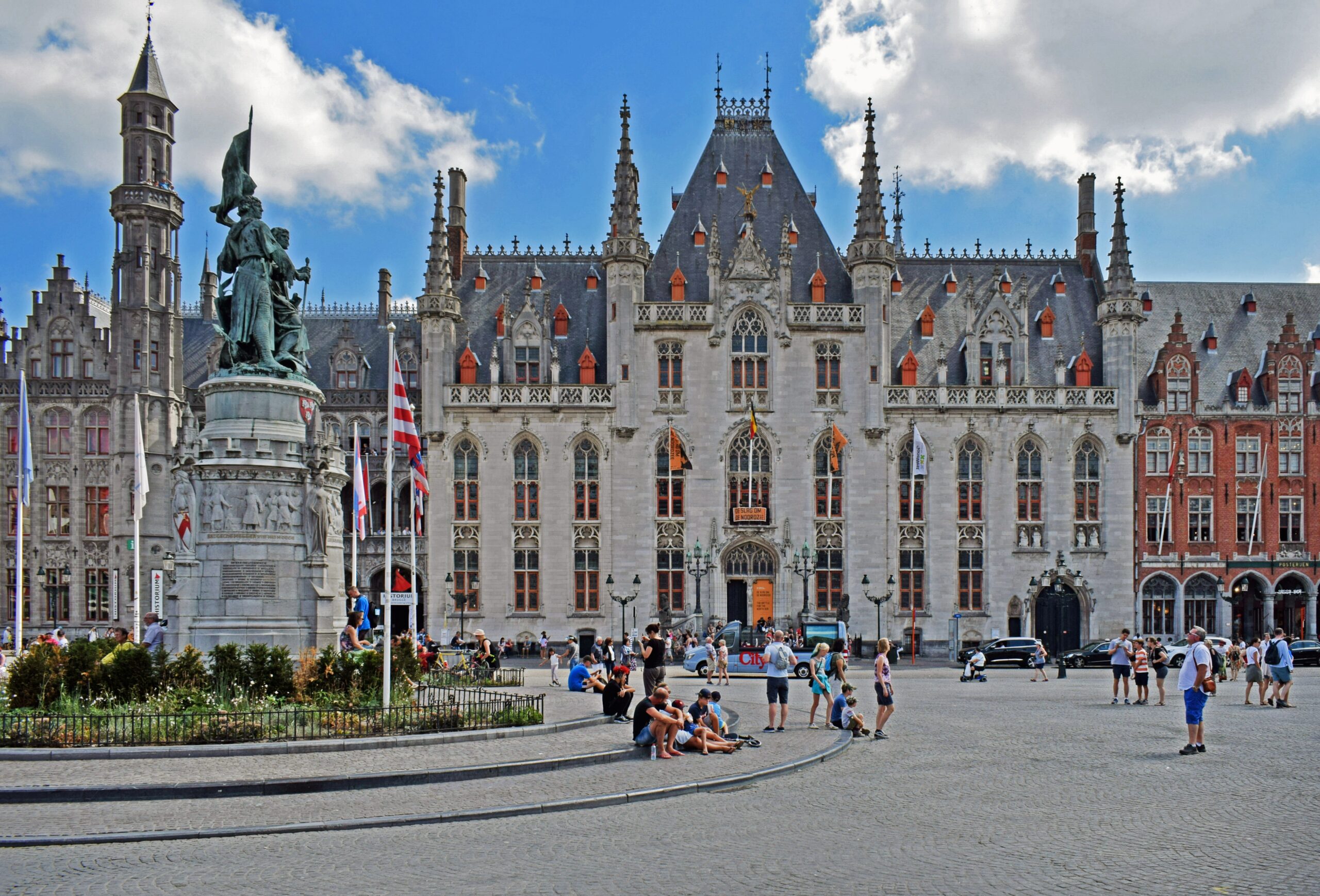 Historisches gotisches Rathaus am zentralen Marktplatz von Brügge in Belgien mit Touristen und Denkmal bei sonnigem Himmel.