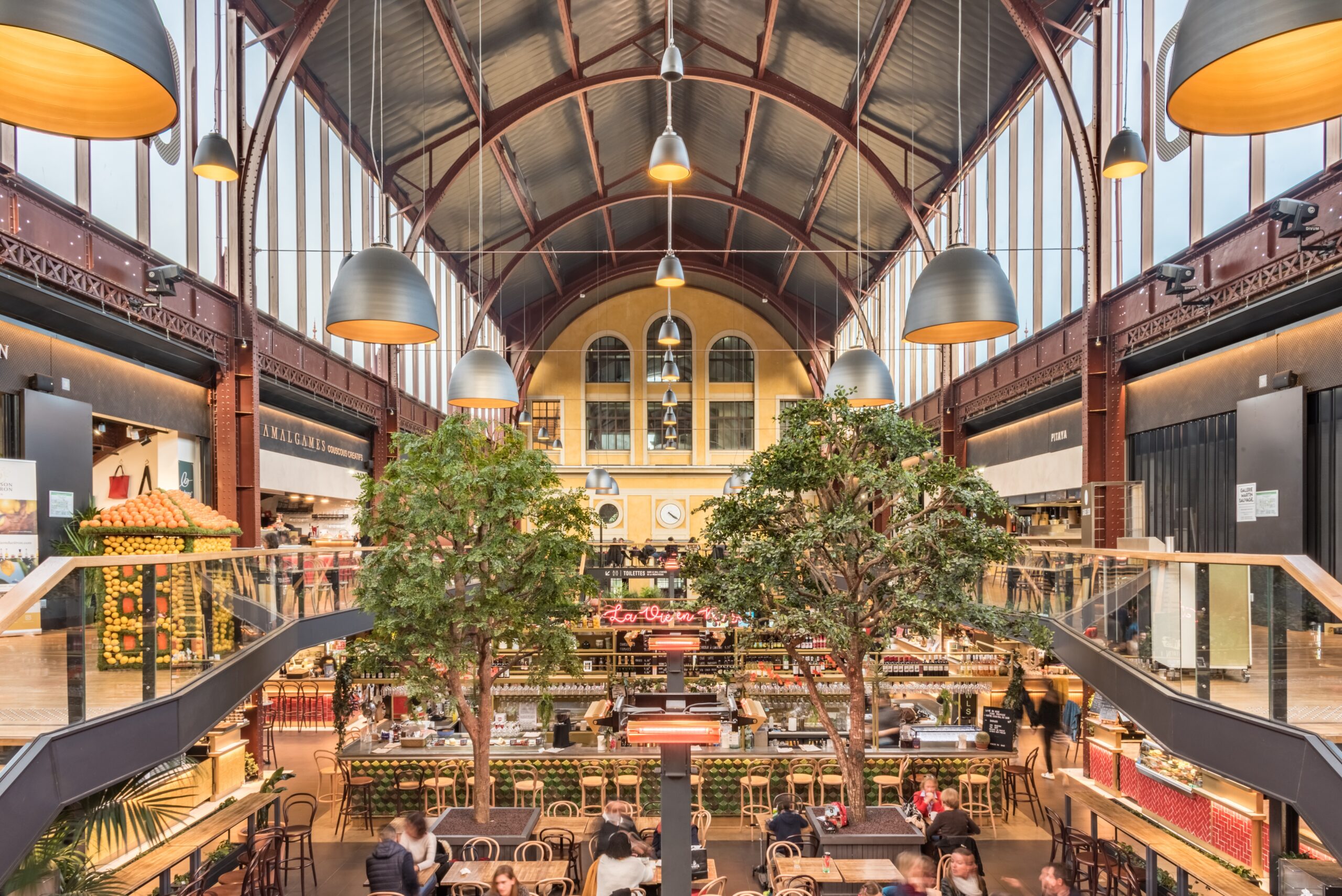 Moderne Markthalle mit Restaurants und Sitzbereichen in einem historischen Industriegebäude in Frankreich mit hohen Stahlträgern, Glasdach und urbanem Ambiente.