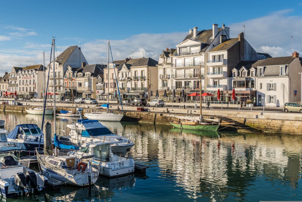 Yachthafen in einer französischen Küstenstadt mit Segelbooten, ruhigem Wasser und traditionellen Häusern entlang der Uferpromenade unter blauem Himmel.